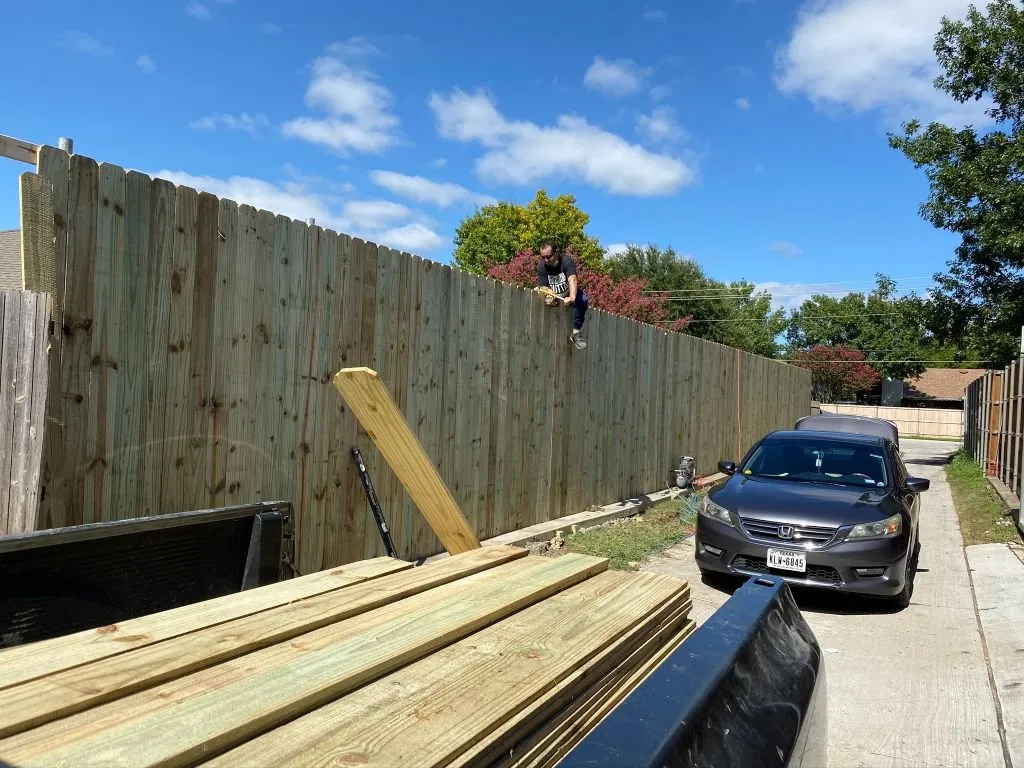 A worker from a care company balances on a wooden fence during landscaping services, preparing the site for landscape maintenance and outdoor lighting installation in Tarrant County for Texas Rangers as part of TX services.